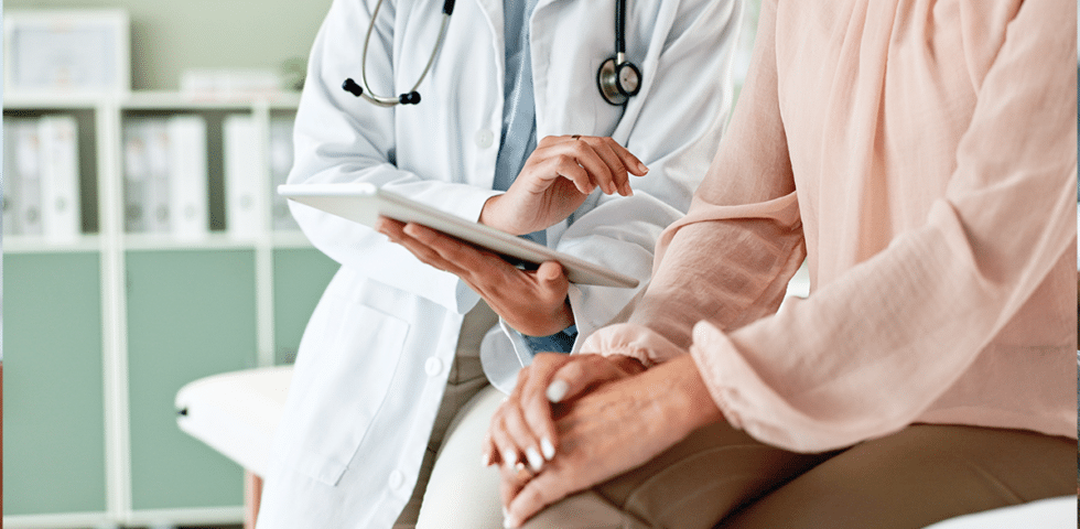 A doctor sitting with a patient and holding a clipboard.