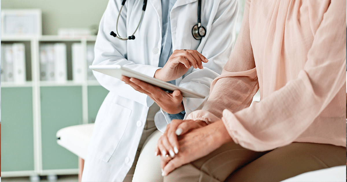 A doctor sitting with a patient and holding a clipboard.